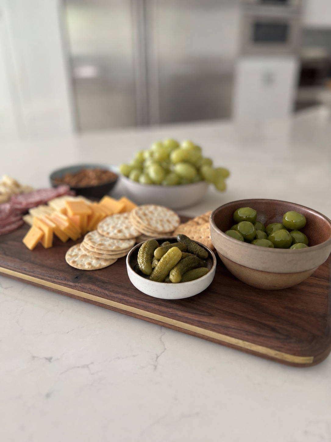 A walnut charcuterie board with a brass inlay, set out with cheese, crackers, olives and pickles